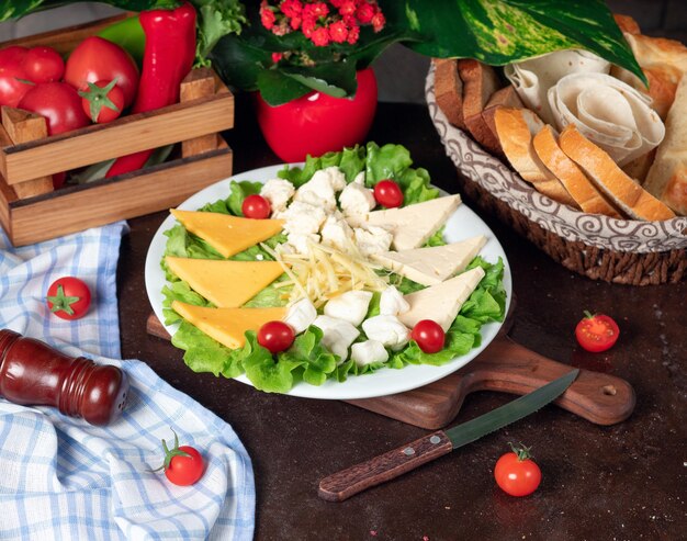 Различные виды сыров расположены на деревянной доске и украшены помидорами черри, листьями салата и свежим хлебом.