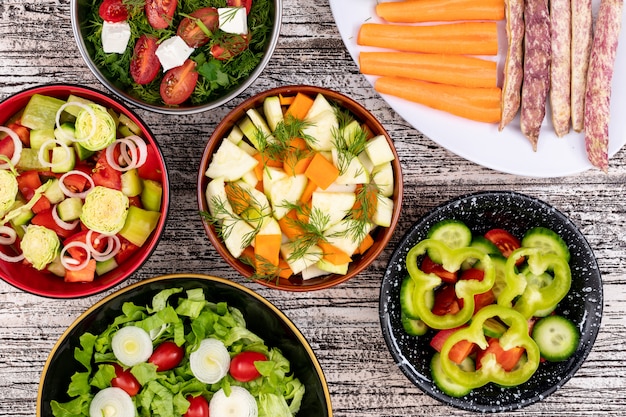 Different vegetable salads in different bowls on white wooden surface sweet pepper beans carrot onion lettuce top view