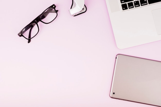 Цифровой планшет; очки и ноутбук на розовом фоне