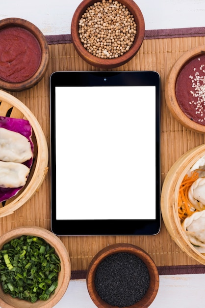 Digital tablet surrounded with steamers; spring onion; sesame seeds and coriander seeds on placemat