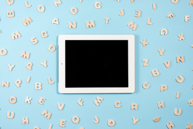Digital tablet with black screen display surrounded with wooden letters on blue background