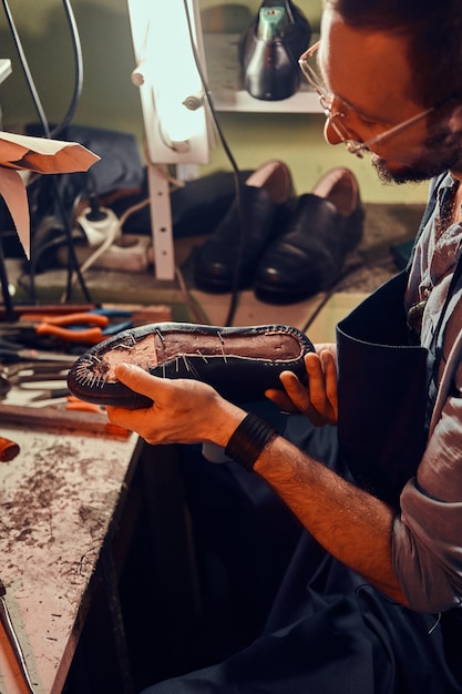 Diligent cobbler in glasses is working with pair of leather shoes at his workplace.