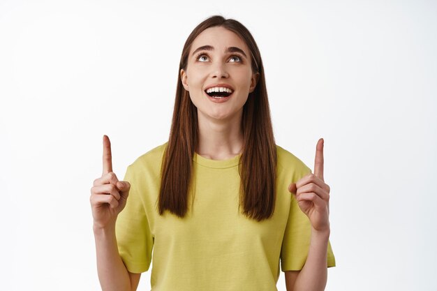 Direction. Portrait of happy young woman pointing and looking up, smiling excited, checking out advertisement, showing promo discount, standing over white background.
