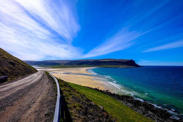 Dirt road near a grassy hill and the sea with a mountain in the distance under a blue sky
