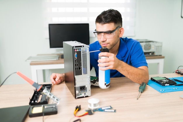 Dirty computer. Attractive young man wearing protection glasses while cleaning inside with compressed air the hardware of a computer