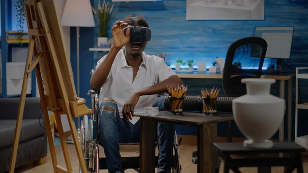 Disabled black artist using vr glasses for virtual inspiration on drawing project at studio. African american young man in wheelchair with modern headset technology for fine art masterpiece