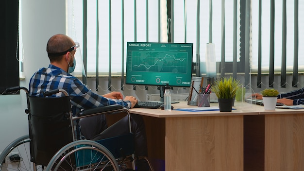 Disabled businessman sitting in wheelchair with protection mask cleaning hands before checking financial data in business modern office. Handicapped freelancer with visor respecting social distance