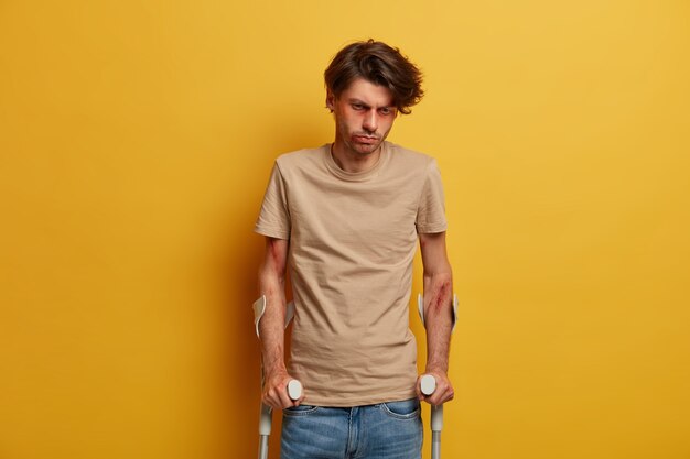 Disabled handicapped bruised man looks sadly down, cannot walk himself for long period of time, recalls dreadful road accident, becomes victim of reckless driving, poses against yellow wall