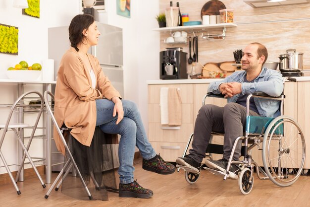 Мужчина-инвалид в инвалидной коляске и жена улыбаются друг другу на кухне. Инвалид, парализованный инвалид, инвалид при ходьбе, интегрирующийся после несчастного случая.