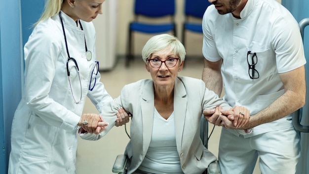 Disabled mature woman getting up from the wheelchair with help of two doctors at clinic