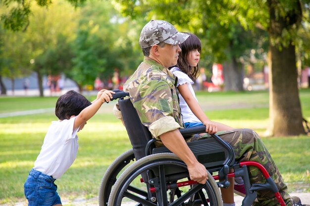 Ветеран-инвалид гуляет с двумя детьми в парке. Девушка сидит на коленях пап, мальчик толкает инвалидную коляску. Ветеран войны или концепция инвалидности