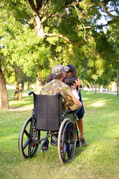 障害者の引退した軍の父親が家に帰り、妻と2人の子供を抱き合ってキスした。背面図。戦争のベテランまたは帰国の概念
