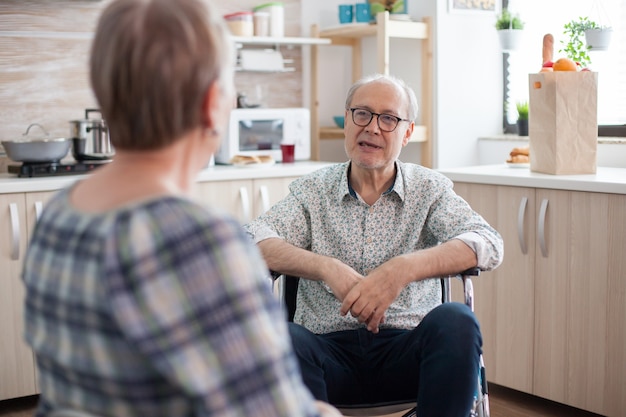 キッチンで妻と話している障害者の年配の男性。キッチンで夫と会話をしているお年寄り。歩行障害のある障害者との生活