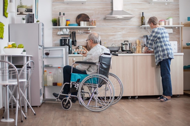 Disabled senior man in wheelchair taking eggs carton from refrigerator for wife in kitchen. Senior woman helping handicapped husband. Living with disabled person with walking disabilities