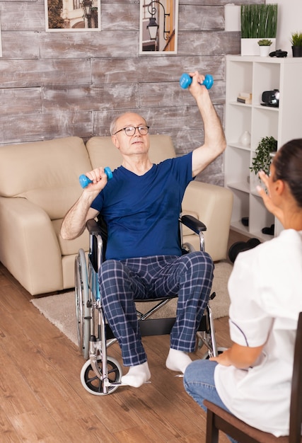 Disabled senior man in wheelchair training with dumbbells during rehabilitation with nurse