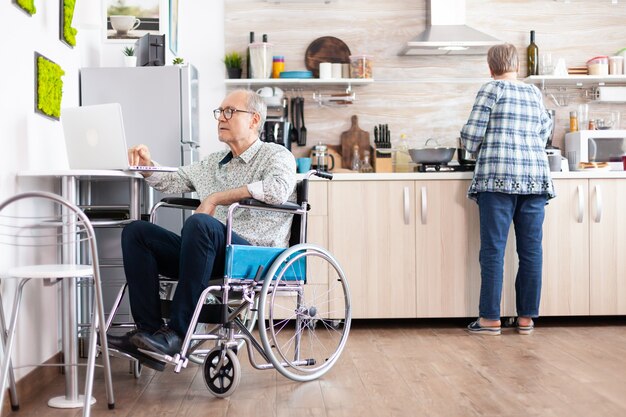 Disabled senior man in wheelchair working from home at laptop in kitchen while wife is cooking breakfast. Handicapped businessman, disabilty entrepreneur paralysis for elderly retired man.