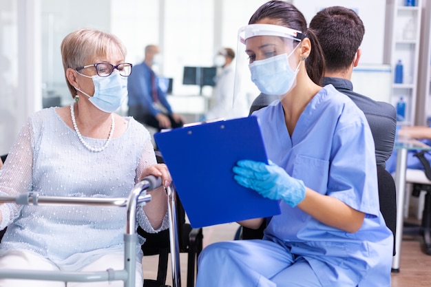 Disabled senior woman having a discussion with medical specialist about treatment in hospital hallway wearing face mask against coronavirus