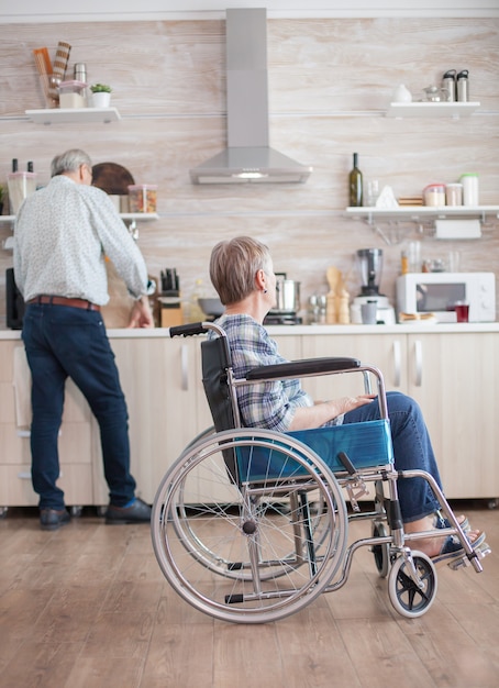 Инвалид старший женщина, сидящая в инвалидной коляске на кухне, глядя через окно. Проживание с инвалидом. Муж помогает жене с инвалидностью. Пожилая пара со счастливым браком.