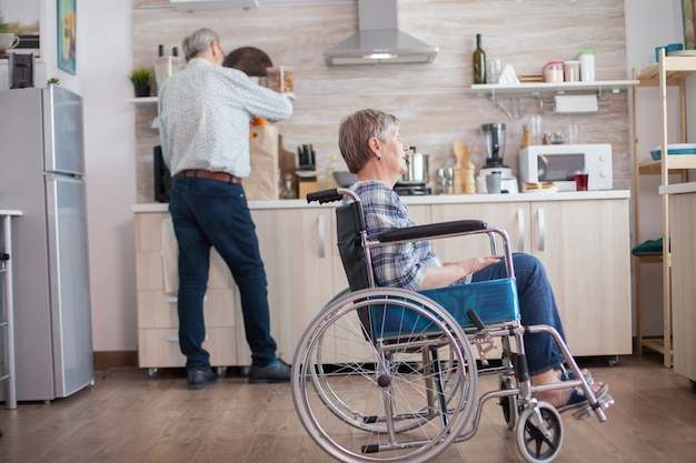 窓越しに見ているキッチンの車椅子に座っている障害者の年配の女性。障害者との生活。夫は障害を持つ妻を助けています。幸せな結婚と年配のカップル。