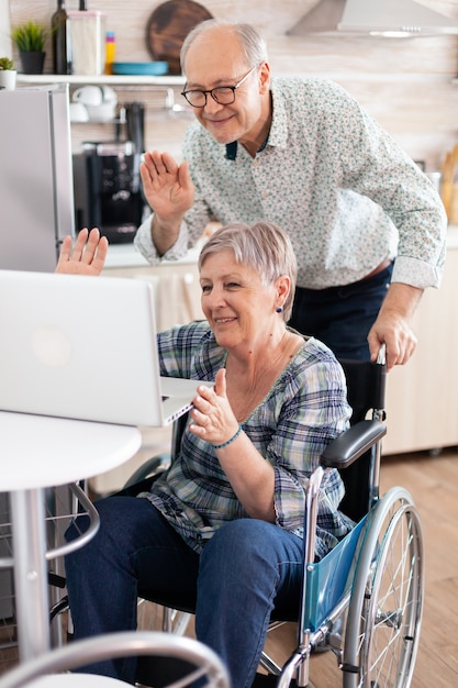 Старшая женщина-инвалид в инвалидной коляске машет рукой во время видеоконференции, сидящей рядом с мужем. парализованная пожилая женщина с ограниченными возможностями и ее муж во время онлайн-звонка с использованием современных коммуникационных технологий.