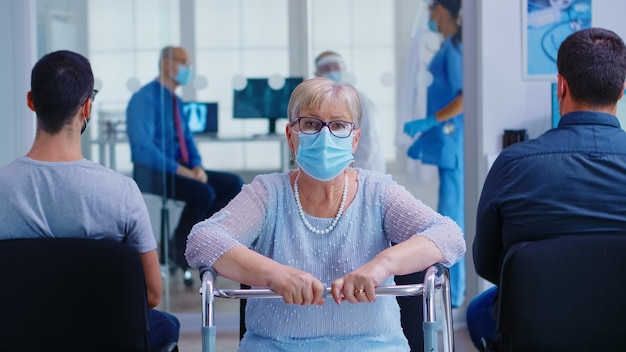 Disabled senior woman with face mask against coronavirus and walking frame looking at camera in hospital waiting area. Nurse assisting doctor during consultation in examination room.