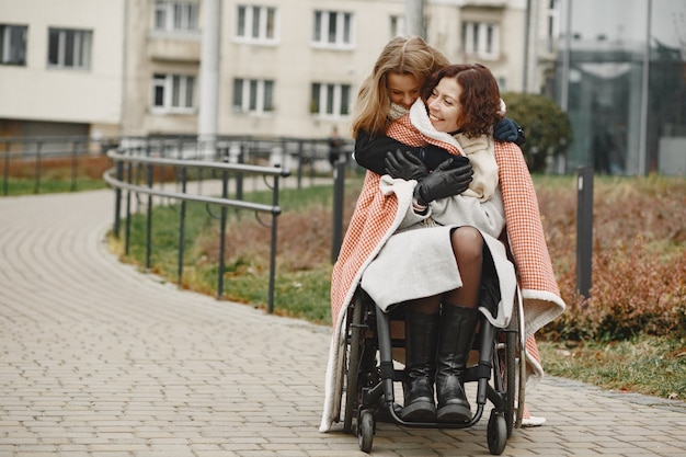 딸과 함께 휠체어에 장애인 여자입니다. 공원에서 밖에 걷는 가족. 어린 소녀는 격자 무늬로 어머니를 덮었습니다.