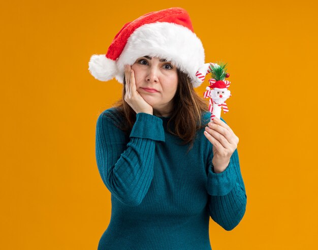 サンタの帽子をかぶった失望した大人の白人女性はあごに手を置き、コピースペースでオレンジ色の背景に分離されたキャンディケインを保持します。