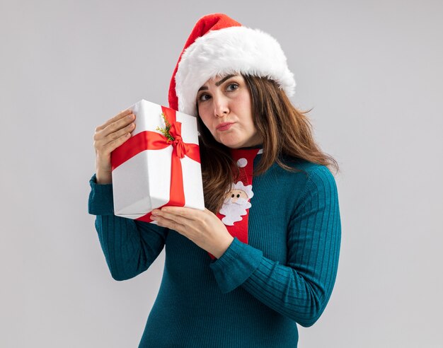 disappointed adult caucasian woman with santa hat and santa tie holding christmas gift box isolated on white background with copy space