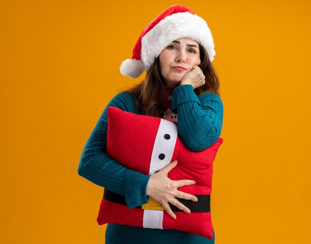 Disappointed adult caucasian woman with santa hat and santa tie puts hand on chin and holds decorated pillow isolated on orange wall with copy space
