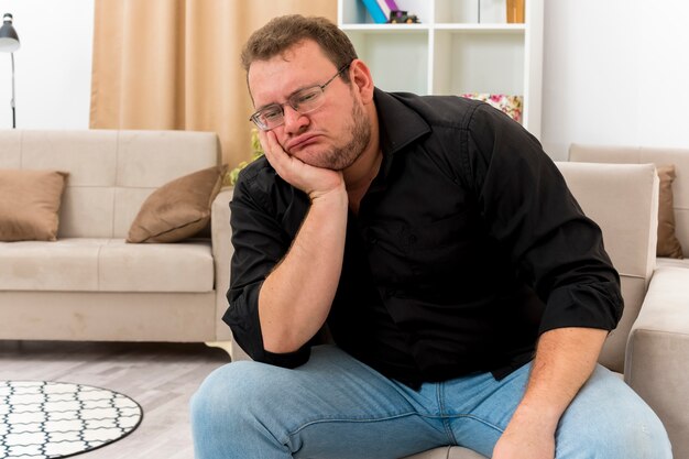 Disappointed adult slavic man in optical glasses sits on armchair holding chin and looking down inside living room