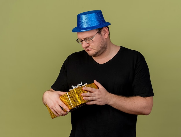 Free photo disappointed adult slavic man in optical glasses wearing blue party hat holds gift box