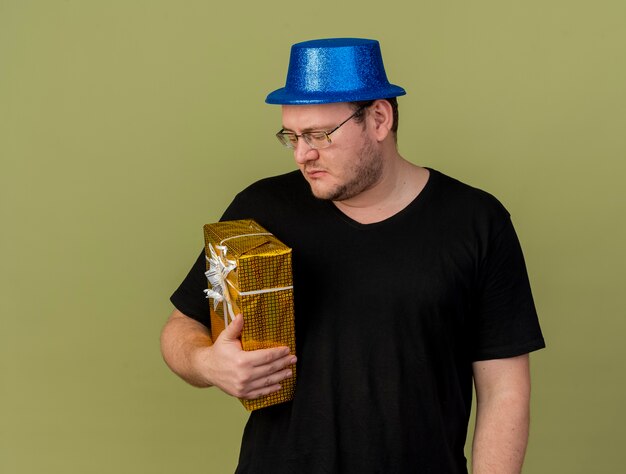 Disappointed adult slavic man in optical glasses wearing blue party hat holds and looks at gift box 