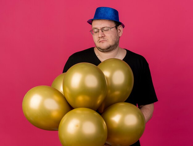 Disappointed adult slavic man in optical glasses wearing blue party hat holds and looks at helium balloons 