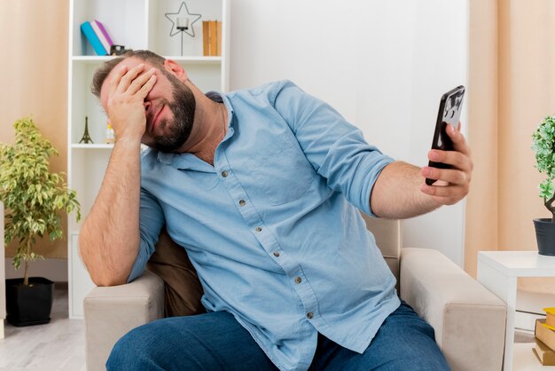 Disappointed adult slavic man sits on armchair putting hand on face and holding phone inside the living room