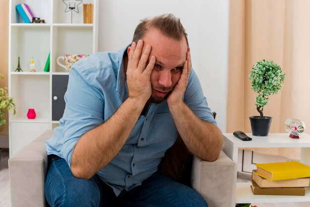 Disappointed adult slavic man sits on armchair putting hands on face looking at side inside the living room