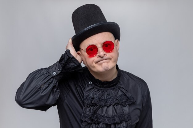 Disappointed adult slavic man with top hat and with sunglasses in black gothic shirt looking at front