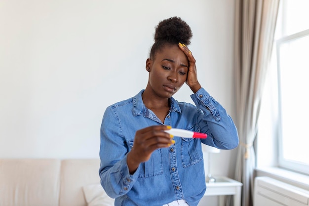 Разочарованная африканская американка, получившая неожиданный результат от набора тестов на беременность Грустная молодая женщина сидит одна дома