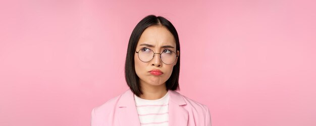 Disappointed asian businesswoman office lady in glasses looking upset at smth unfair sulking standing over pink background