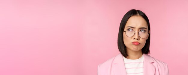 ピンクの背景の上に立っているsmth不公平なやめなさいに動揺して見ている眼鏡の失望したアジアの実業家のオフィスの女性