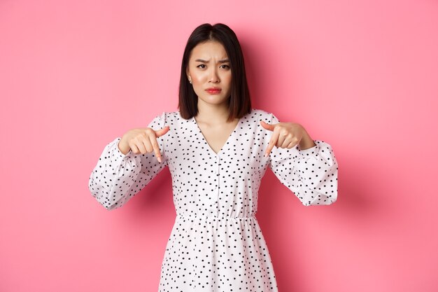 실망한 아시아 여성 모델은 프로모션 제안에 손가락을 아래로 가리키고, 인상을 찡그리며 카메라를 쳐다보고, 분홍 배경 위에 서 있습니다.