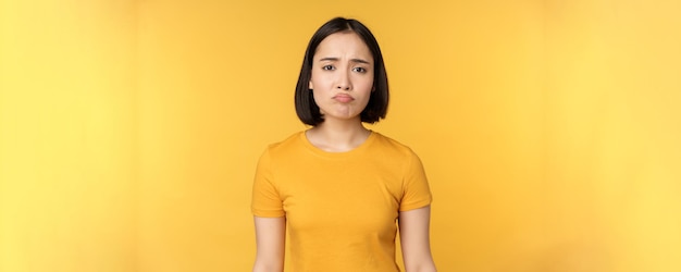 失望したアジアの女の子は、白い背景の上に黄色のTシャツを着て、気分を害しているように見えます。