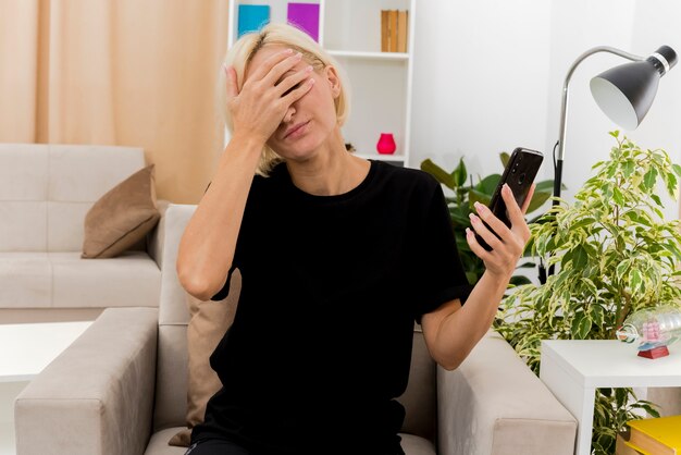 Disappointed beautiful blonde russian woman sits on armchair putting hand on face holding phone inside the living room