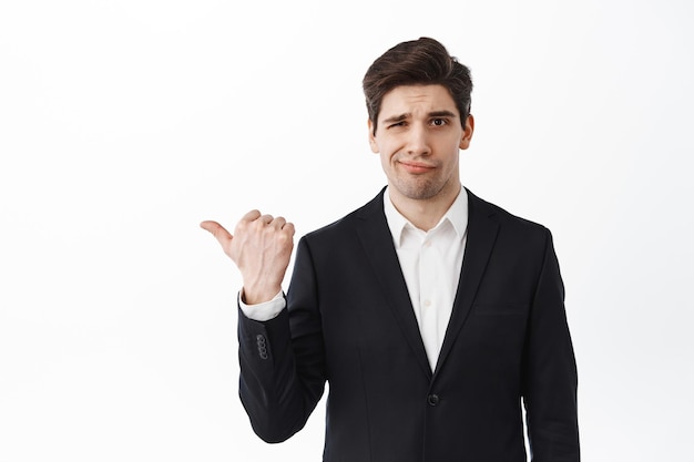 Disappointed business man frowning, pointing left at bad work, displeased or doubtful about advertisement, standing in suit against white background