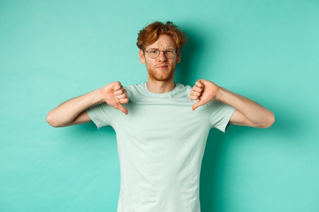 Disappointed caucasian guy with red messy hair and glasses showing thumbs down, dislike something, frowning upset and displeased, standing over mint background.