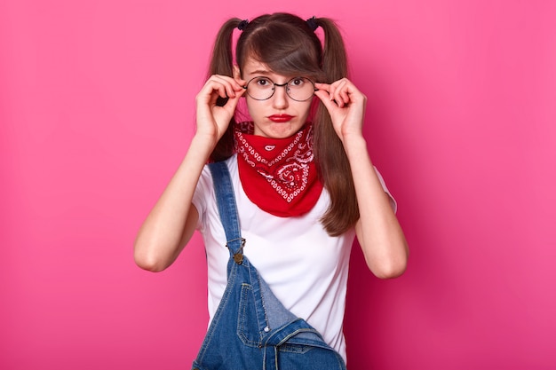 Disappointed charismatic young lady with bangs and long pigtails has frown face, touches her eyeglasses, choosing comfortable position