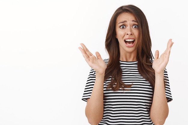 Disappointed concerned attractive brunette female with freckles in striped t-shirt raising hands in despair and disappointment, cringe and grimacing embarassed, look upset camera white wall