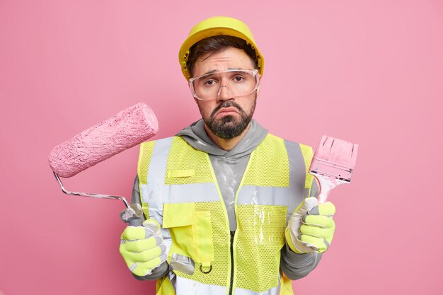 Disappointed construction worker holds painting roller and brush restores paints walls of building wears protective helmet uniform and safety glasses 