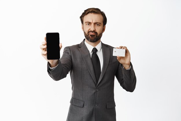 Disappointed corporate man showing smth upsetting on mobile phone screen and holding credit card furrow eyebrows and grimacing displeased white background