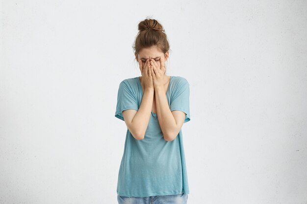 Разочарованная удрученная женщина с пучком волос в голубой футболке, закрывающей лицо руками, уставшими и измученными. Отчаявшаяся женщина в депрессии, пряча плачущее лицо руками