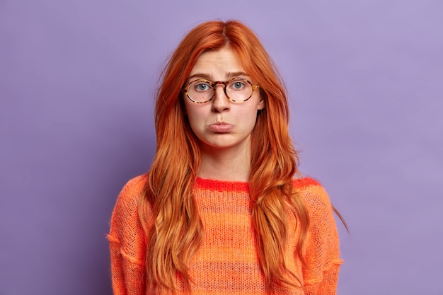 Disappointed displeased redhead woman purses lips being sad as someone hurt her feelings complains about something wears optical glasses and jumper.